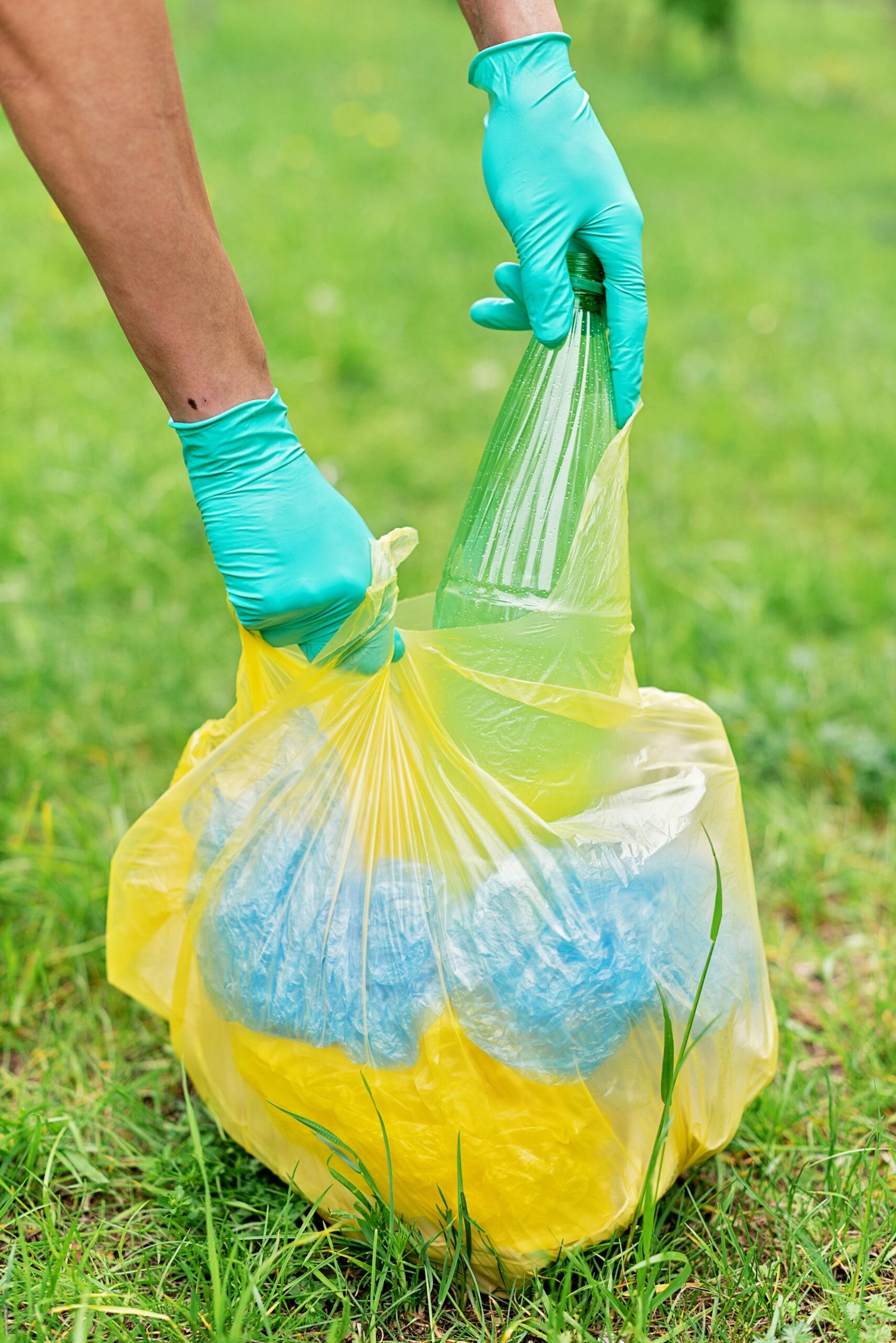 Person wearing gloves collecting recyclable bottles in a yellow plastic bag outdoors.